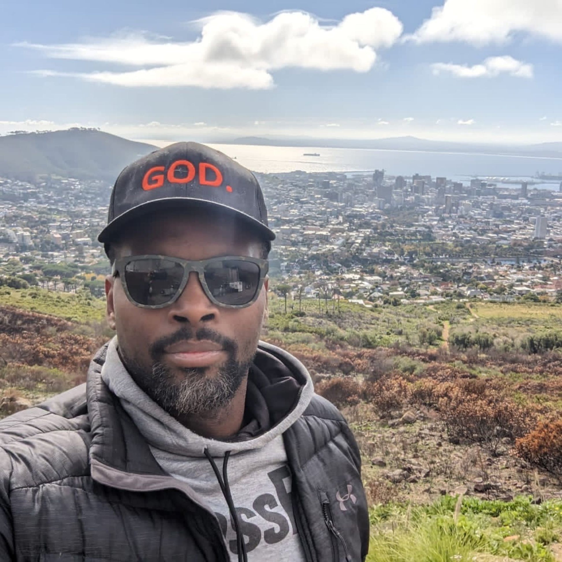 Curtis Harkness wearing a black GOD. trucker cap overlooking a coastal city and ocean—D&B Originals.